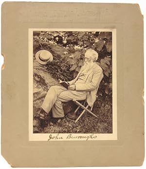 An excellent photograph of John Burroughs, seated, reading, next to a large rock and tree with hi...