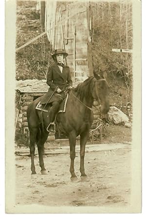 [Real Photo Postcard--Natty Woman Astride on Horseback in Front of Log Shed and Partial Wall]