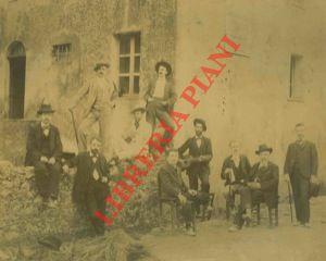 Gruppo di persone in piedi e sedute (uno con chitarra) in posa attorno alla casa natale di Giosuè...
