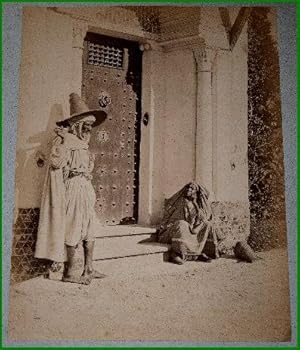 Bild des Verkufers fr Grande photographie anciennes originale (Vers 1880). Tirage albumin d'poque mont sur carton bleu-vert reprsentant un homme arabe et une femme discutant devant une porte de maison. zum Verkauf von JOIE DE LIRE