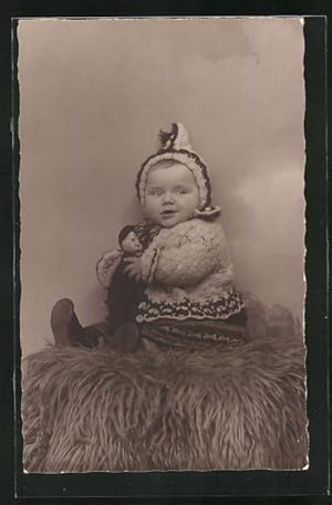 Foto-Ansichtskarte Süsses Baby in winterlicher Kleidung mit Puppe auf Fell sitzend