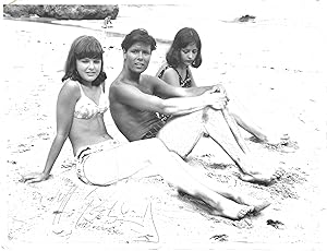 Bild des Verkufers fr [Sir Cliff Richard, popular British singer and film actor.] Autograph Signature to black and white print of publicity photograph of him in swimming trunks, seated on a beach between two girls in bikinis. zum Verkauf von Richard M. Ford Ltd